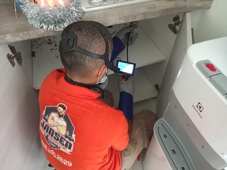 Pest control technician inspecting under-sink cabinet using camera tool, wearing gloves and headlamp, with Jansen branding visible on red shirt